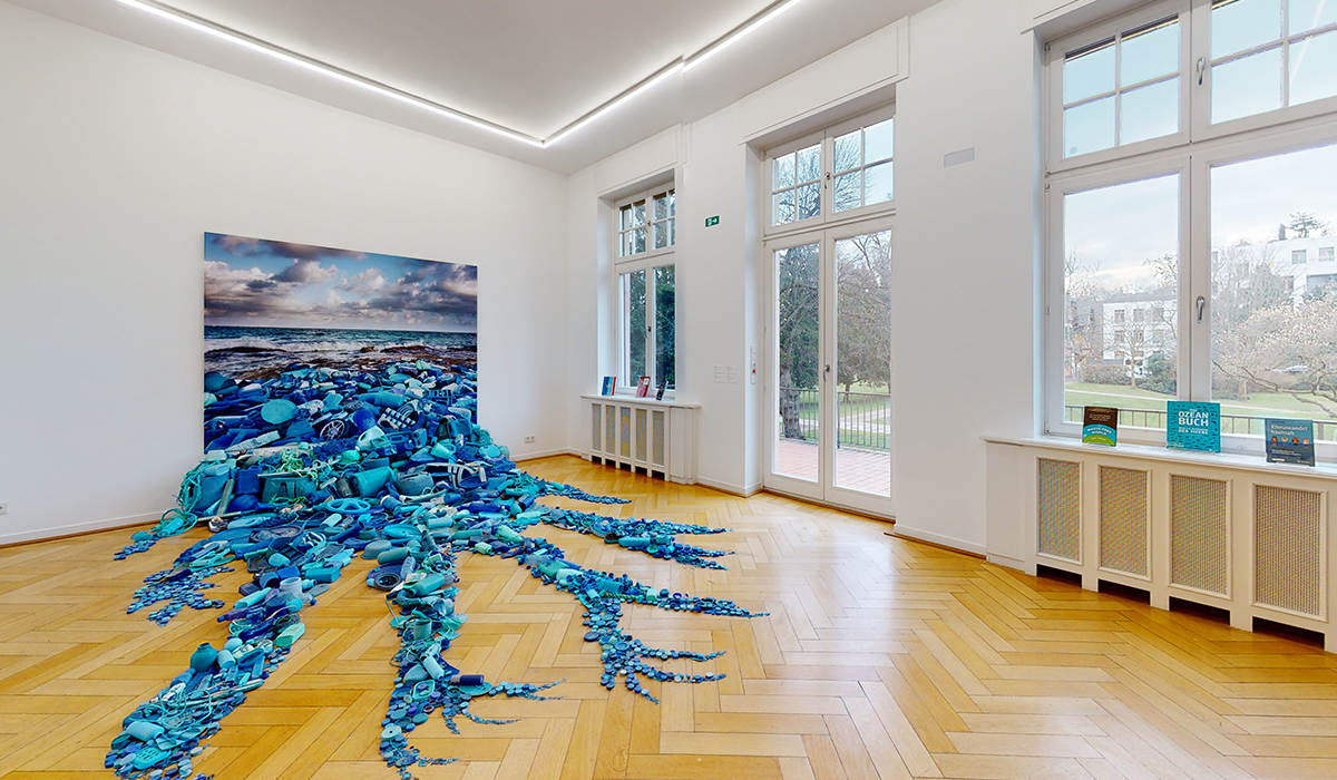 Kunstinstallation in einem modernen Ausstellungsraum mit Parkettboden und großen Fenstern. Im Vordergrund blaue Plastikobjekte, die aus einem Foto mit Meeresmotiv auf dem Boden herausfließen – thematisiert Meeresverschmutzung durch Plastik.